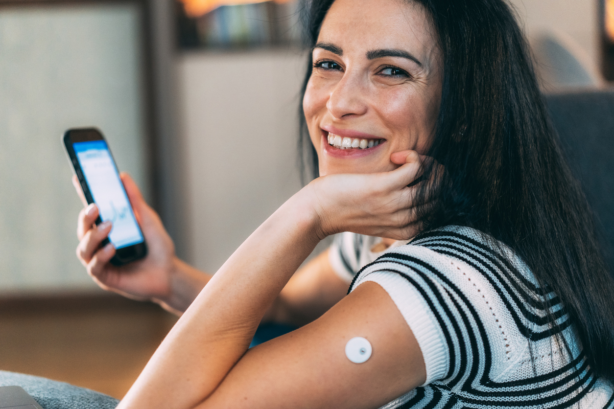 А woman smiling while holding her smartphone, with a sensor attached to her arm, likely for measuring blood sugar levels. She appears to be reviewing data or graphs from a health-related app on her phone. This scene captures the intersection of technology and personal health management, highlighting the importance of monitoring health metrics in a modern lifestyle. The woman's expression conveys positivity and engagement, making it an ideal representation of proactive health awareness.
